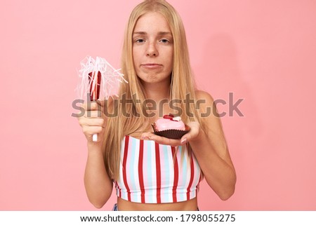 Horizontal image of unhappy young Caucasian woman with loose fair hair and freckles posing with piece of cake, eating sweet dessert, holding horn, being lonely and upset on her birthday party