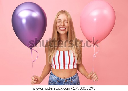 Studio image of cute happy young European female wearing braces and crop top closing eyes while making wish on her birthday, enjoying party with friends, smiling, holding two shiny helium balloons
