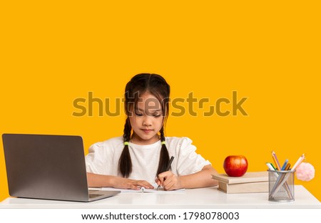 Asian Schoolgirl Doing Homework Writing Sitting At Laptop Over Yellow Studio Background. Learning Classes, School Knowledge Concept