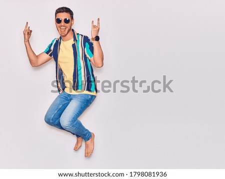 Young handsome hispanic man wearing casual clothes and sunglasses smiling happy. Jumping with smile on face doing horns sign with fingers over isolated white background