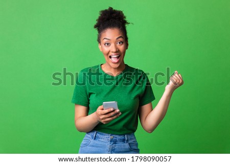 Big Luck. Emotional african girl holding cell phone shouting and gesturing yes standing on green wall