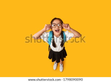 School Nerd. Funny Asian Schoolgirl Wearing Eyeglasses Smiling To Camera Posing Over Yellow Studio Background. High-Angle Shot