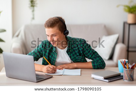 Joyful teen guy writing at notebook and looking at laptop screen, having lesson online, home interior, free space Royalty-Free Stock Photo #1798090105