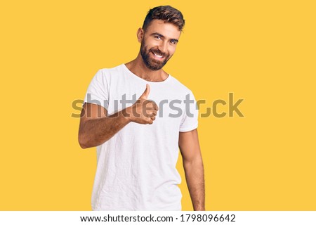 Young hispanic man wearing casual white tshirt doing happy thumbs up gesture with hand. approving expression looking at the camera showing success. 
