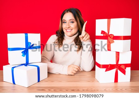 Young beautiful caucasian woman sitting on the table with presents smiling with an idea or question pointing finger up with happy face, number one 
