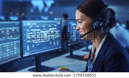 Beautiful Female Data Scientist Works on Personal Computer Wearing a Headset in Big Infrastructure Control and Monitoring Room. Woman Engineer in a Call Center Office Room with Colleagues. Royalty-Free Stock Photo #1798108855