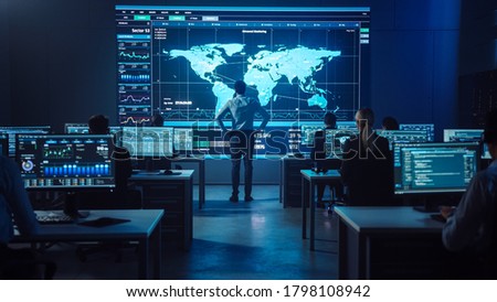 Project manager Stands Next to Big Screen with Neural Network. Team of Professional Computer Data Science Engineers Work on Desktops in a Dark System Control and Monitoring Telecommunications Office. Royalty-Free Stock Photo #1798108942
