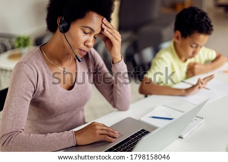 Stay at home mother using laptop and holding her head in pain while her son is doing homework.  Royalty-Free Stock Photo #1798158046