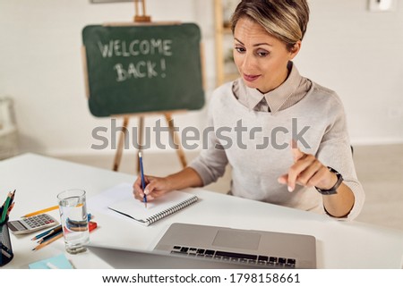 Female teacher taking notes while having video call with her students over laptop at home. 