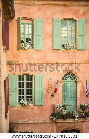 Vertical close-up picture of town hall in Roussillion, Provence, France. Old historical building with orange walls and green windows. Provencal and french flags near balcony door. Tourism destination.
