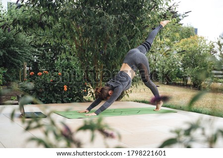 Active young woman in three legged downward dog pose on exercise mat with laptop in backyard. High quality photo