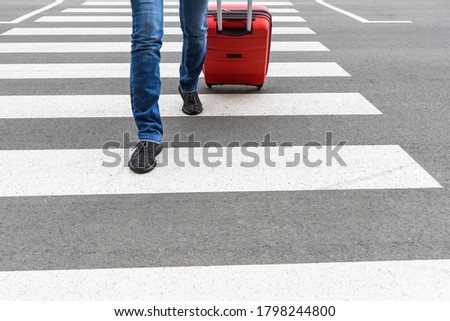 The close up view of woman legs in blue jeans and red rolling bag on the pedestrian crossing. The background is unfocused with art noise.