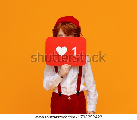 Ginger boy in trendy outfit covering face with board with like symbol and looking at camera while advertising social media against yellow background
