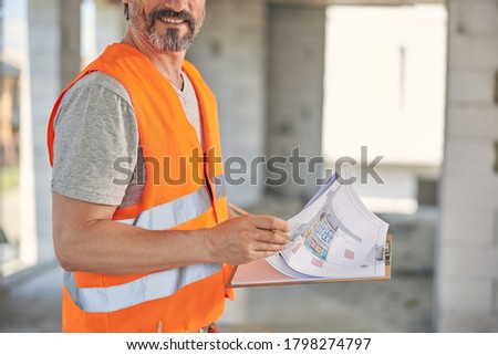 Cropped photo of a bearded male worker holding a clipboard with blueprints in his hands