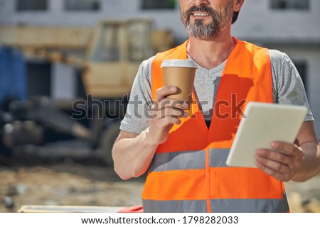 Cropped photo of a builder in a reflective waistcoat holding a paper cup in his hand