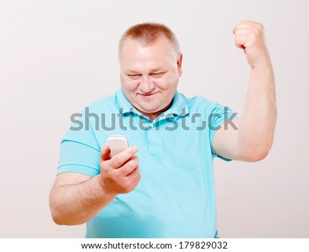 Middle aged man in blue shirt celebrating success with mobile phone over white background
