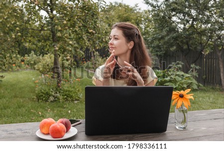 Young woman in a light T-shirt remotely working on a laptop sitting at a wooden table on a background of green garden and looking at the side