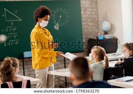 Happy black teacher and her students wearing protective face mask in the classroom. Teacher is giving them their test results.  Royalty-Free Stock Photo #1798363270