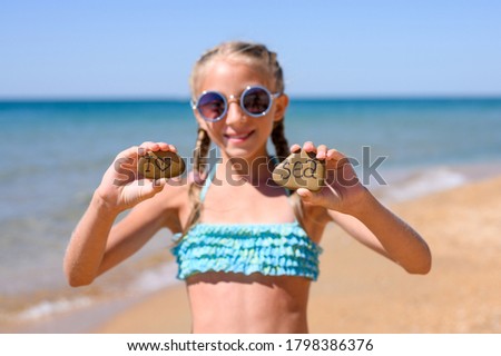Joyful girl in sunglasses on the beach stands and shows two stones with the words I love the sea. Focus on the pebbles with the inscription.