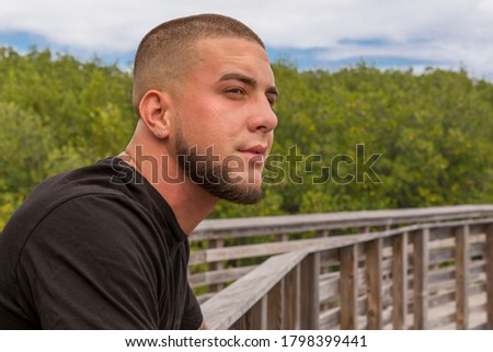 Outdoors leaning on the boardwalk rail, he looks across the way with a staring gaze. A scared face tough guy with a military buzz cut leans on the wood rail with a focused look. Royalty-Free Stock Photo #1798399441