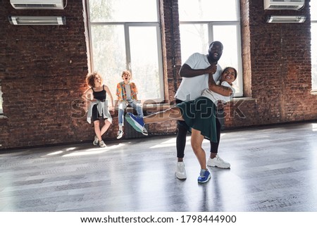 Smiling young couple learning modern dances in studio with their friends on the background