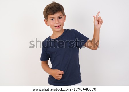 Smiling Beautiful kid boy wearing casual t-shirt standing over isolated white background pointing up and looking at the camera 