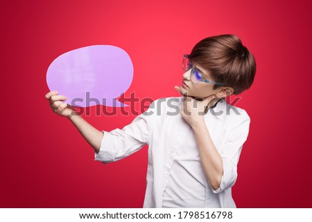 Caucasian boy with red hair and eyeglasses is advertising something looking at the blank space touching chin on a red studio wall