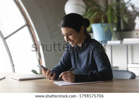 Smiling young indian businesswoman filling paper document form, web surfing important information online on smartphone. Distracted from paperwork happy mixed race employee chatting in social network.