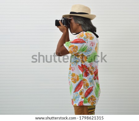 Elderly traveler asian man wearing summer shirt, straw hat, sunglasses and brown shorts holding camera in hands standing over white wall, Business summer holiday concept