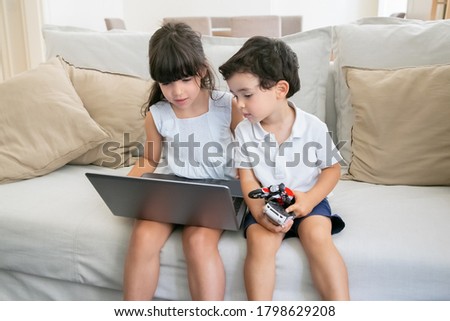 Cute boy and girl sitting on couch at home and using laptop, watching video or movie. Front view, medium shot. Kids leisure time or communication concept
