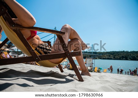 Selective focus on the legs of an attractive young woman lying on a yellow couch on a beach steam. The beach is sandy and is located on the river bank and behind is a bridge.