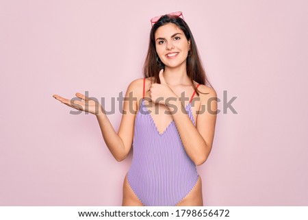Young beautiful fashion girl wearing swimwear swimsuit and sunglasses over pink background Showing palm hand and doing ok gesture with thumbs up, smiling happy and cheerful