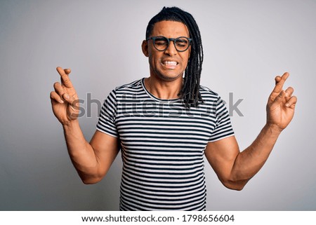 Young handsome african american man with dreadlocks wearing striped t-shirt and glasses gesturing finger crossed smiling with hope and eyes closed. Luck and superstitious concept.