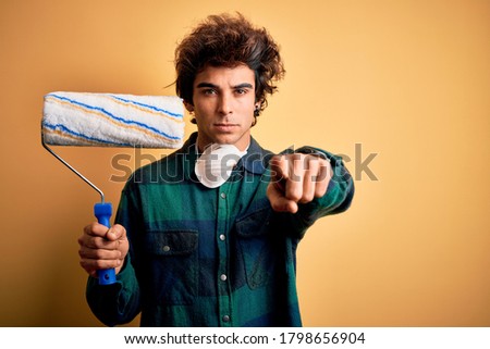 Young handsome painter man holding roller standing over isolated yellow background pointing with finger to the camera and to you, hand sign, positive and confident gesture from the front