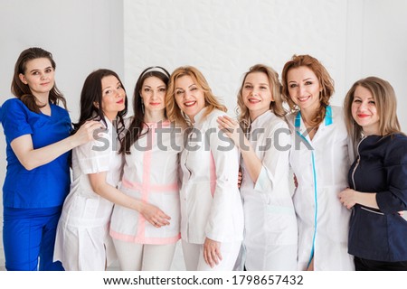 Beautiful young female doctors in white uniforms posing against the background of a white wall. Women hugging as a sign of team unity