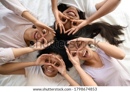 Group of cheerful diverse girls best friends lying in bed in pajamas making binoculars with fingers looking at camera top view. Bachelorette party, sleepover hanging out, friendship having fun concept