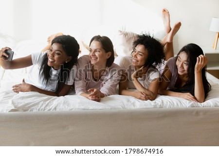 African ethnicity girl having fun with best friends enjoy sleepover in pajamas lying in bed. Womans holding smart phone, taking selfie. Girlfriends make photo in memory of bachelorette party