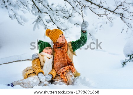 Two cute little cheerful children are playing with snow among snowdrifts. Children sit in the snow under snow-covered tree. Winter activities for children. Knitted scarf, hat, sweater, woolen coat.