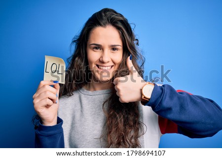 Young beautiful woman with curly hair holding reminder paper with lol message happy with big smile doing ok sign, thumb up with fingers, excellent sign Royalty-Free Stock Photo #1798694107