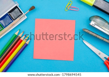 Pink sheet of paper with copy space on a blue background with colored pencils, scissors, paints, stapler, paper clips, top view.