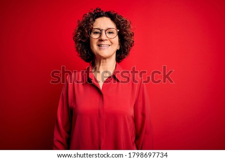 Middle age beautiful curly hair woman wearing casual shirt and glasses over red background with a happy and cool smile on face. Lucky person.