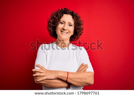 Middle age beautiful curly hair woman wearing casual t-shirt over isolated red background happy face smiling with crossed arms looking at the camera. Positive person.