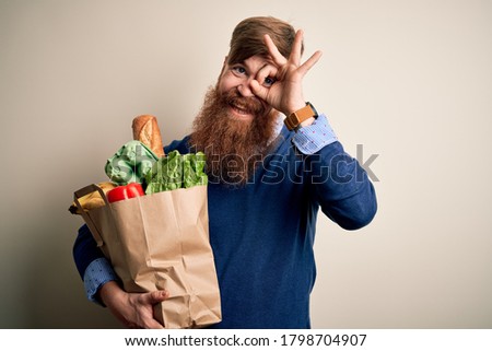 Redhead Irish man with beard holding fresh groceries from supermarkt over isolated background with happy face smiling doing ok sign with hand on eye looking through fingers