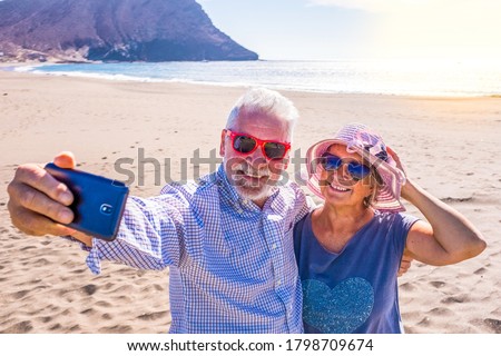 couple of mature people or pensioners enjoying their vacations and summer time together on the sand of the beach with the sea or ocean at the background - two retired senior taking a selfie smiling Royalty-Free Stock Photo #1798709674