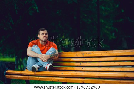 Adult man resting on bench. Adult male in red t shirt and jeans sitting on bench and looking away while resting in park on summer day