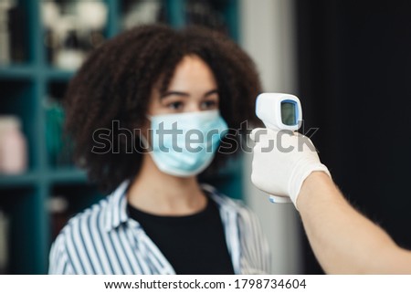 Body temperature check. Young black woman in medical mask getting thermal screening with contactless thermometer, close up