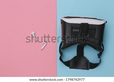 VR headset and wireless headphones on blue-pink pastel background. Modern gadgets. Top view