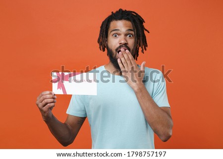 Shocked young african american man wearing blue casual t-shirt posing isolated on orange wall background studio portrait. People lifestyle concept. Hold gift certificate covering mouth with hand