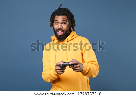 Excited young african american man in casual yellow streetwear hoodie isolated on blue wall background studio portrait. People sincere emotions lifestyle concept. Play game with joystick