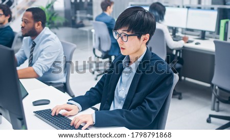 Young Professional Japanese Stock Broker Work on His Desktop Computer. Diverse and Motivated Business People Work in Modern Open Office.